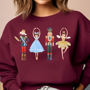 Op de afbeelding: Een bordeauxrood sweatshirt met een feestelijk ontwerp met een Notenkraker, een ballerina en een muizenkoning. De personages zijn geïllustreerd in een grillige stijl met felle kleuren en ingewikkelde details.