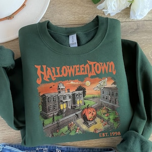 May include: A dark green sweatshirt with a graphic print of a spooky town with a large pumpkin in front of a building. The text "HALLOWEEN TOWN EST. 1998" is printed on the bottom of the graphic.