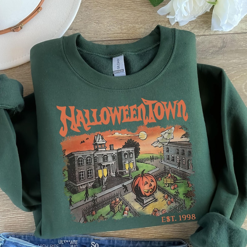 May include: A dark green sweatshirt with a graphic print of a spooky town with a large pumpkin in front of a building. The text "HALLOWEEN TOWN EST. 1998" is printed on the bottom of the graphic.