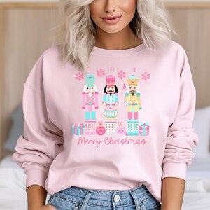 Op de afbeelding: Roze trui met een kerstontwerp met drie notenkrakers in roze, blauw en geel met een roze, blauw en wit sneeuwvlokkenpatroon. De tekst "Merry Christmas" is onder de figuren gedrukt.