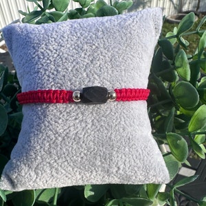 May include: A red macrame bracelet with a black rectangular center bead and two small gold-colored beads. The bracelet is displayed on a gray, textured pillow, with green foliage in the background.