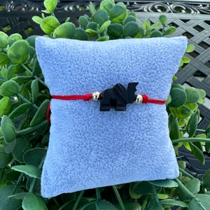 May include: A red string bracelet with a black elephant charm and silver beads. The bracelet is displayed on a light blue fuzzy surface.