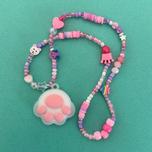 Op de afbeelding: Een ketting met kralen en een pootafdruk hanger. De ketting heeft roze, paarse en witte kralen, waaronder hartvormige en stervormige accenten. De pootafdruk hanger is wit met roze voetzolen.
