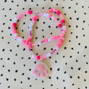 Op de afbeelding: Een roze en witte kralenketting met een roze pootprint bedel. De ketting ligt op een witte achtergrond met zwarte sterren.