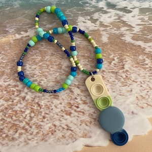 Puede incluir: Un collar de cuentas azules, verdes y blancas con un colgante de juguete pop-it. El colgante está hecho de silicona y tiene tres círculos de diferentes tamaños. El collar está sobre un fondo de playa de arena.