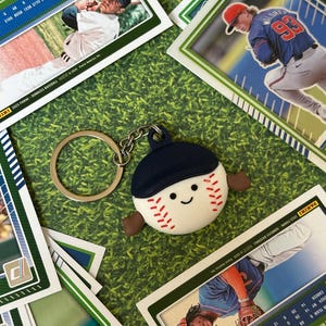 Könnte beinhalten: Ein Baseball-Schlüsselanhänger mit einem lächelnden Baseball-Charakter, der eine marineblaue Kappe trägt, umgeben von Baseballkarten auf einer grünen Oberfläche. Der Schlüsselanhänger hat einen silbernen Ring und eine Kette.