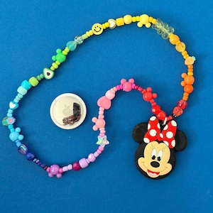 Op de afbeelding: Een kleurrijke kralenketting met een Minnie Mouse-hanger en een zilveren ronde tag. De ketting heeft een regenboog aan kralen, waaronder smiley-gezichten, harten en sterren. De Minnie Mouse-hanger heeft een rode strik met witte stippen.