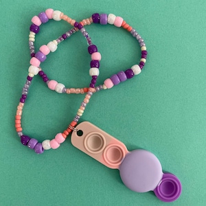May include: A purple, pink, and white beaded necklace with a multi-colored fidget toy attached. The fidget toy has three circles that can be pushed in and out.
