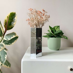 Puede incluir: Un jarrón de espejo plateado con un diseño geométrico, lleno de flores secas en tonos blancos y rosados. El jarrón está sobre una mesa blanca.