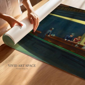 May include: A rolled-up art print featuring a vintage painting of a diner scene with people at the counter. The print is being unrolled on a wooden surface. The text "VIVID ART SPACE" and "Printable Vintage Art" are visible.
