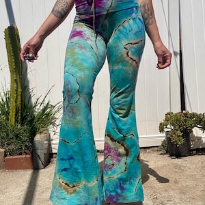 Può includere: Un paio di pantaloni a zampa d'elefante blu, marrone e viola con stampa tie-dye. I pantaloni hanno vita alta e gamba larga.