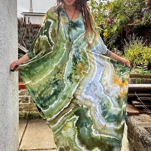 May include: A flowing, tie-dye maxi dress in shades of green, white, and gold. The dress features a V-neckline, short sleeves, and a gathered waist. The fabric has a marbled effect, creating a unique, artistic look. The dress is worn outdoors.