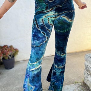 Puede incluir: Pantalones acampanados de terciopelo azul y turquesa con un diseño veteado. Los pantalones de cintura alta tienen una amplia campana en la parte inferior. La tela tiene un aspecto suave y lujoso, con vetas azul oscuro y negro.