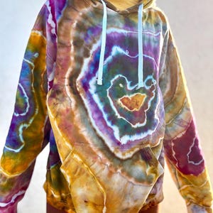 Op de afbeelding: Een tie-dye hoodie in tinten paars, geel en bruin. De hoodie heeft een voorzak en een capuchon met trekkoord. Het tie-dye patroon is cirkelvormig, met een hartvorm in het midden. De hoodie is gemaakt van een zacht materiaal.