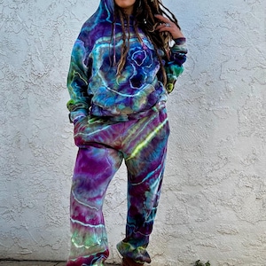 May include: A tie-dye hoodie and matching sweatpants set in shades of purple, blue, and green. The hooded sweatshirt has a drawstring and the sweatpants have a relaxed fit. The outfit is worn with brown boots.