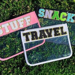 May include: Three clear plastic zippered pouches with different colored fabric tops. The pouches have gold glitter lettering that reads "Stuff", "Travel", and "Snacks".