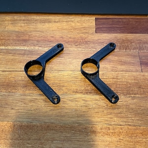 Puede incluir: Dos soportes de plástico negro impresos en 3D con un agujero circular en el centro de cada brazo.