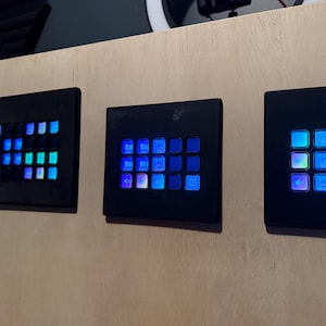 May include: Three black electronic control panels with illuminated blue and green square buttons are mounted on a light wood surface. The panels vary in size and button arrangement, creating a modern, tech-focused aesthetic.