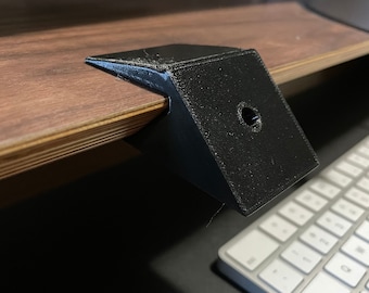 May include: Black 3D printed desk mount with a hole in the centre. The mount is attached to a wooden desk.