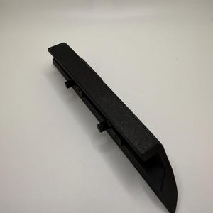 Puede incluir: Objeto negro angular con una superficie superior texturizada. El objeto tiene un extremo afilado y varios salientes pequeños a lo largo del lado. El artículo parece estar hecho de un material negro mate.