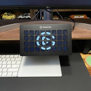 May include: A black Stream Deck with a blue and white logo and 32 customizable buttons. The Stream Deck is sitting on a desk with a white keyboard and a white mouse pad.