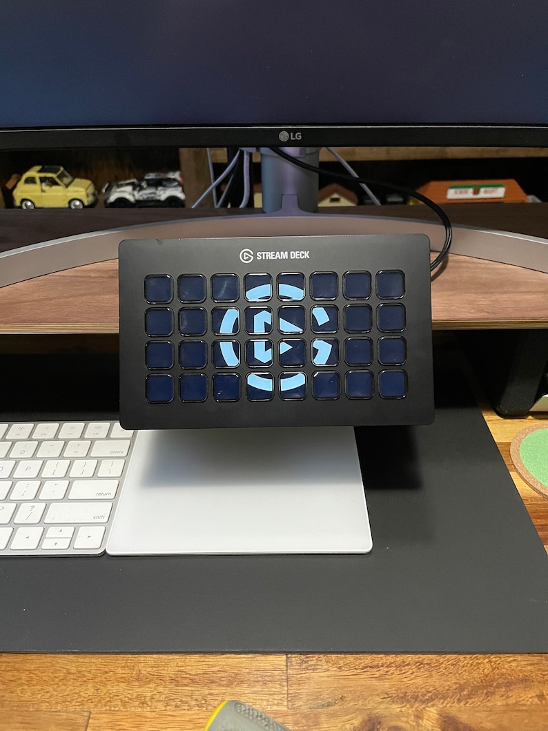 May include: A black Stream Deck with a blue and white logo and 32 customizable buttons. The Stream Deck is sitting on a desk with a white keyboard and a white mouse pad.