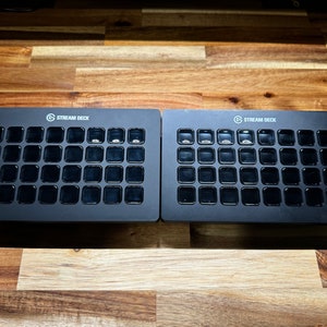 May include: Two black Stream Deck devices with a grid of 15 buttons each. The buttons are black with a clear plastic cover. The devices are on a wooden surface.