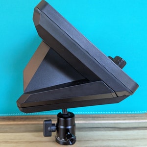 Pode incluir: Um dispositivo eletrônico preto, possivelmente um monitor ou painel de controle, inclinado sobre uma cabeça de tripé preta. O dispositivo tem um design geométrico com uma superfície texturizada e um botão. A cabeça do tripé é montada em uma superfície de madeira contra um fundo turquesa.