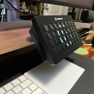 May include: A black Stream Deck with 15 customizable buttons, each with a white LCD screen displaying a different icon. The Stream Deck is mounted on a desk with a white keyboard and mouse pad.