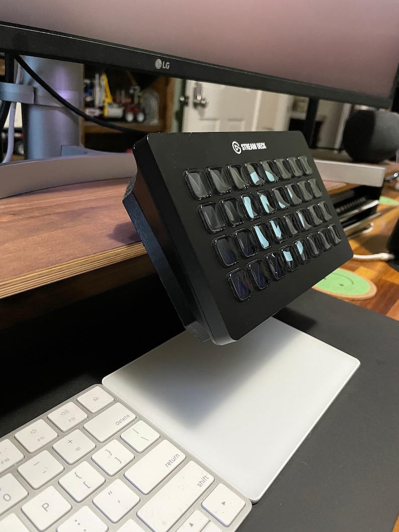 May include: A black Stream Deck with 15 customizable buttons, each with a white LCD screen displaying a different icon. The Stream Deck is mounted on a desk with a white keyboard and mouse pad.