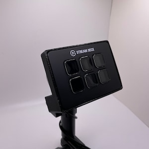 May include: A black Stream Deck mini with six buttons, mounted on a black adjustable arm. The Stream Deck is a device used for live streaming and content creation.