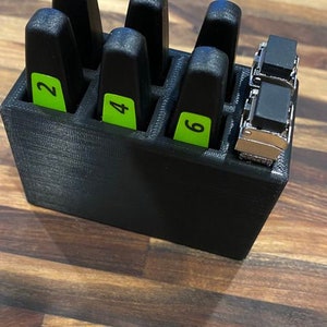 May include: Black plastic storage box with six slots for holding small items. Each slot has a green label with a number, 2, 4, and 6 are visible. The box is sitting on a wooden surface.