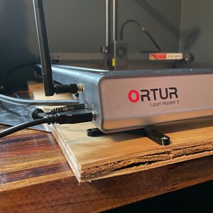May include: A close-up of a silver laser engraver with the text "ORTUR Laser Master 3" on the front. The engraver is sitting on a wooden surface with black cables connected to it.
