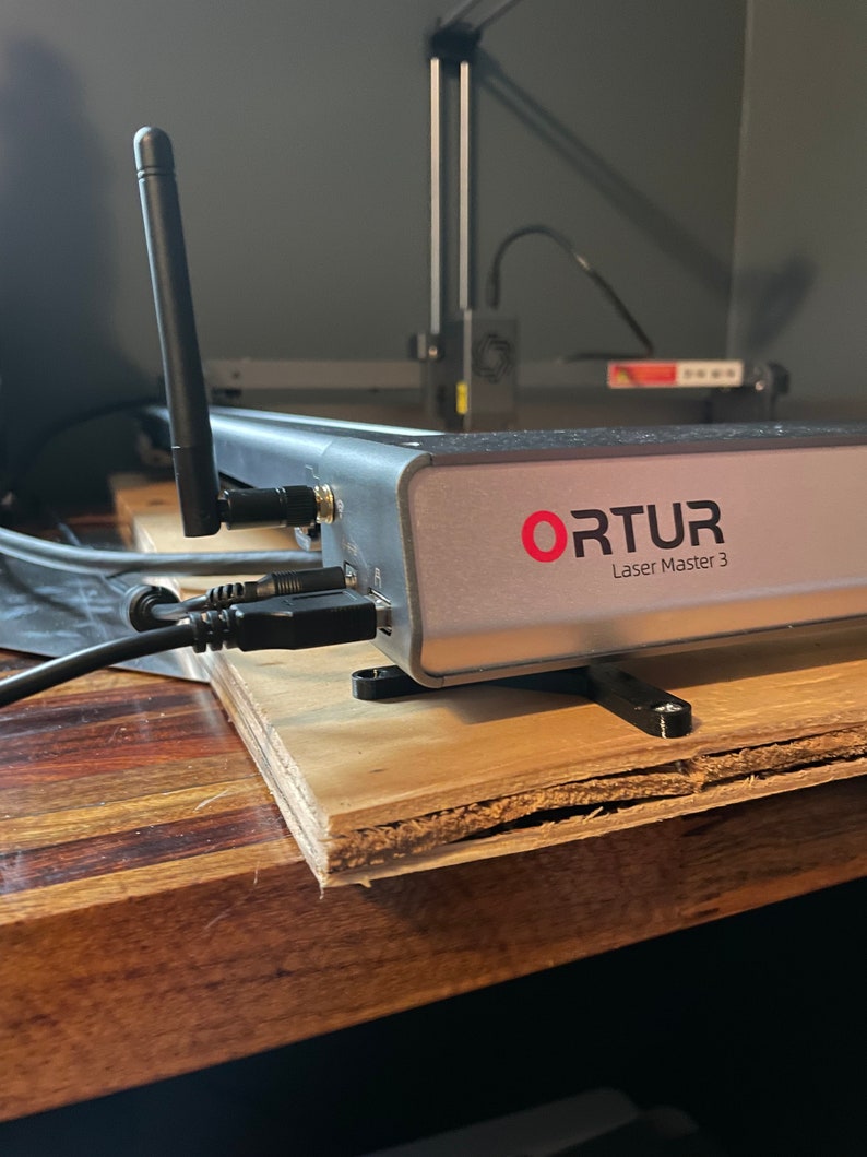 May include: A close-up of a silver laser engraver with the text "ORTUR Laser Master 3" on the front. The engraver is sitting on a wooden surface with black cables connected to it.