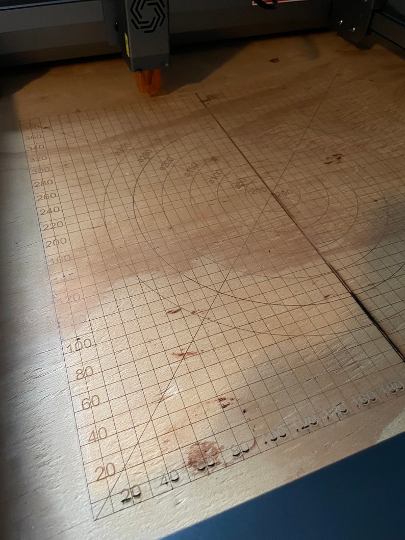 May include: A close-up of a light brown wooden surface with a grid pattern and concentric circles. The grid is marked with numbers from 20 to 340 and the circles are marked with numbers from 100 to 350.