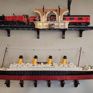 May include: A red and black toy train set with a station and a passenger car. The train is on a shelf above a white and black toy ship with yellow lifeboats.