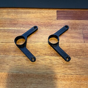May include: Two black plastic 3D printed brackets with a circular hole in the center of each arm.