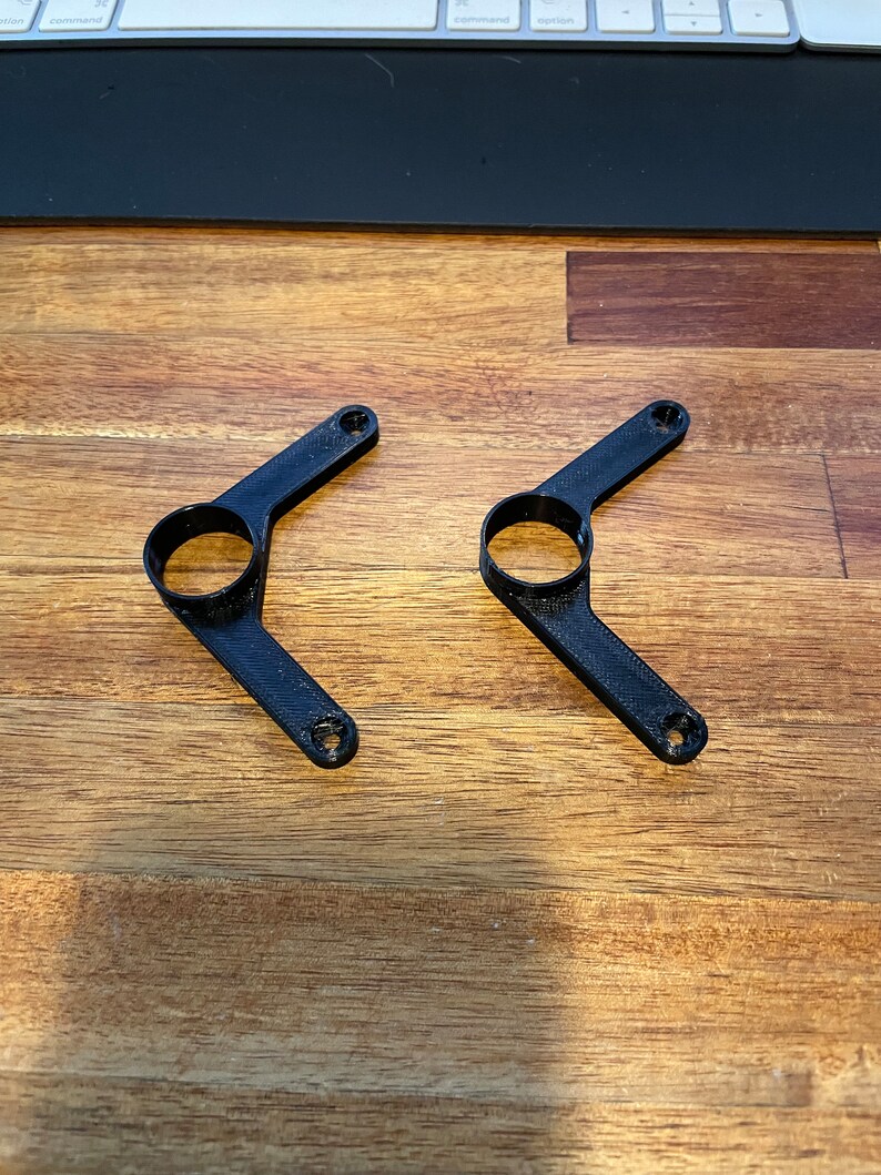 May include: Two black plastic 3D printed brackets with a circular hole in the center of each arm.
