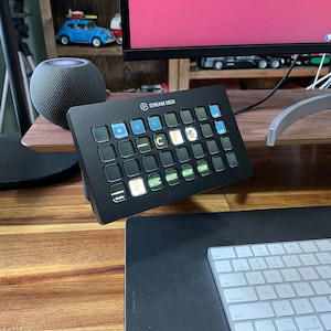 May include: A black Stream Deck with a grid of 15 customizable buttons. The buttons are illuminated with various colors and icons. The Stream Deck is sitting on a wooden desk with a keyboard and mouse mat.