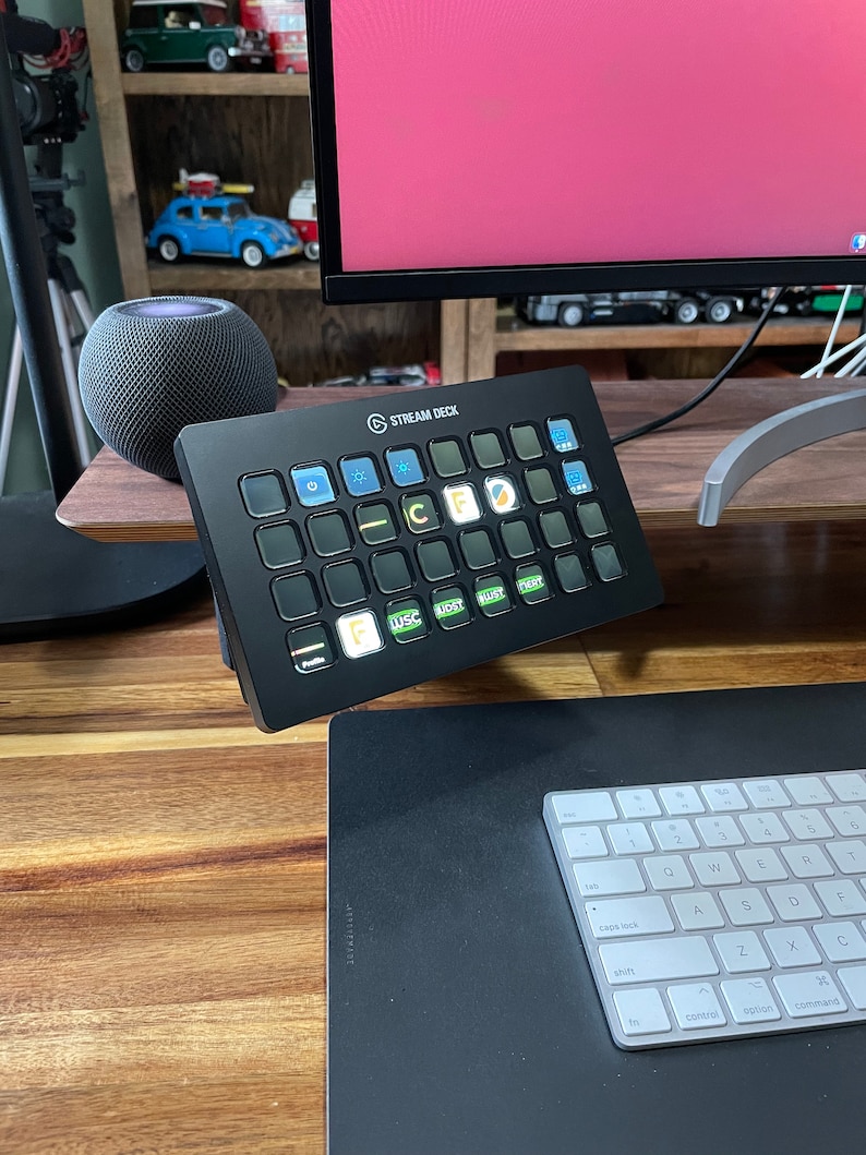 May include: A black Stream Deck with a grid of 15 customizable buttons. The buttons are illuminated with various colors and icons. The Stream Deck is sitting on a wooden desk with a keyboard and mouse mat.