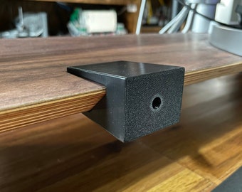 May include: A black plastic monitor stand riser with a small hole in the top. The riser is sitting on a wooden surface.