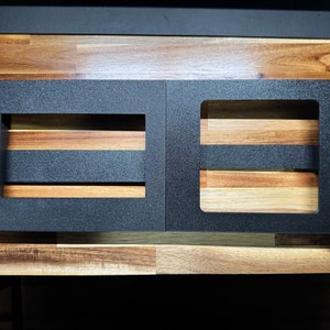May include: Two black metal plates with rectangular cutouts, designed to hold electronic equipment. The plates are on a wooden surface.