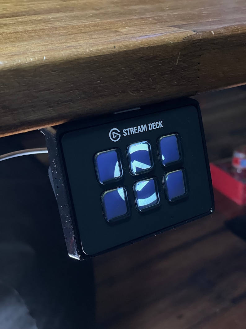 May include: A black Stream Deck with six buttons, each with a different icon. The buttons are illuminated with a blue light. The Stream Deck is mounted to a wooden surface.