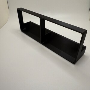 May include: A black plastic shelf with two compartments. The shelf is designed to hold small items, such as pens, pencils, or other office supplies.
