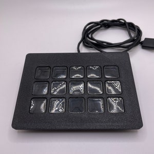 May include: A black, rectangular, electronic device with a grid of 20 clear, square buttons. The device is connected to a black cable.