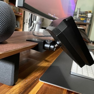 May include: A black monitor stand with a small monitor attached to it. The stand is mounted to a wooden desk with a black mouse pad and a white keyboard on the desk.