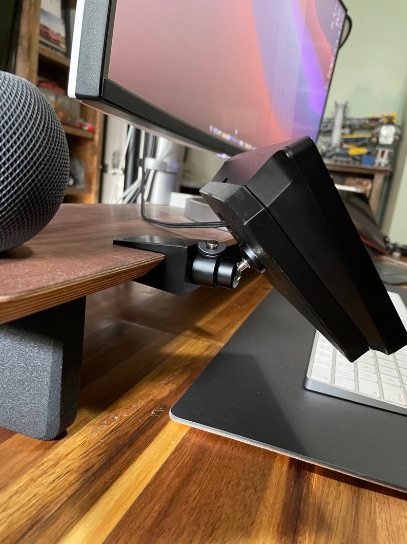 May include: A black monitor stand with a small monitor attached to it. The stand is mounted to a wooden desk with a black mouse pad and a white keyboard on the desk.