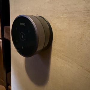 May include: A black and silver BenQ device mounted on a light brown wooden surface. The device is circular with a black face and silver trim. It has a power button and other symbols.