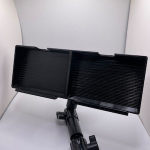 May include: A black phone holder with two adjustable slots, mounted on a black adjustable arm with a knob. The holder is designed to hold two phones or other devices.