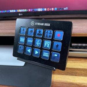 May include: A black Stream Deck with a variety of buttons, including a microphone, camera, person, group of people, red dot, globe, left arrow, right arrow, play button, mute all, sound FX, light bulb, count down, and a button with a file icon. The Stream Deck is on a wooden surface.